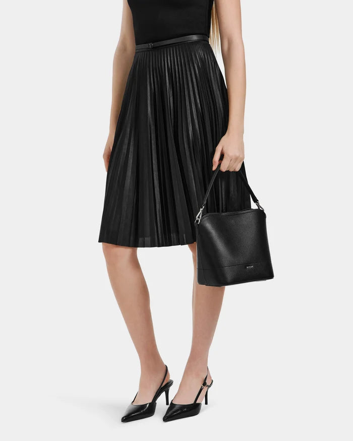 A woman in a black sleeveless top, chic heels, and a pleated midi skirt holds the Rudsak JANE - 8525029 Black leather handbag at her side against a plain white background.
