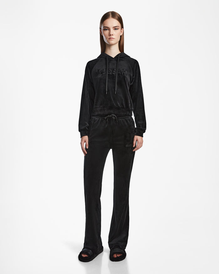 A woman faces forward wearing Rudsak’s JODIE - L523501 Black velour hoodie and matching velvet pull-on pants with an elastic waistband and straight-leg fit, paired with black open-toe slides against a plain white background.