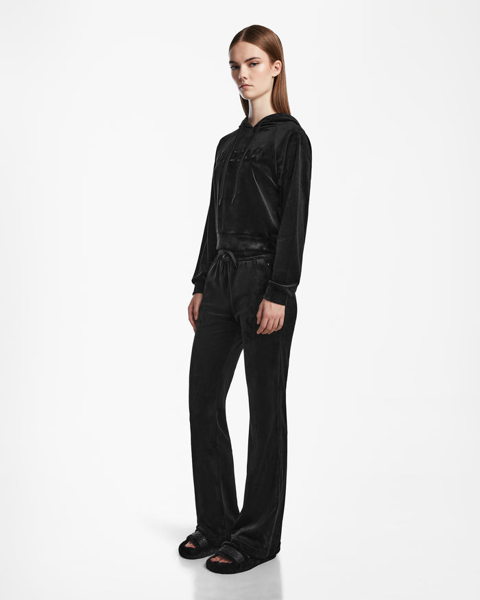 A person with long straight brown hair and a neutral expression stands against a white background, wearing the Rudsak JODIE - L523501 Black velour tracksuit with a hooded sweatshirt and straight-leg pants, paired with black sandals.