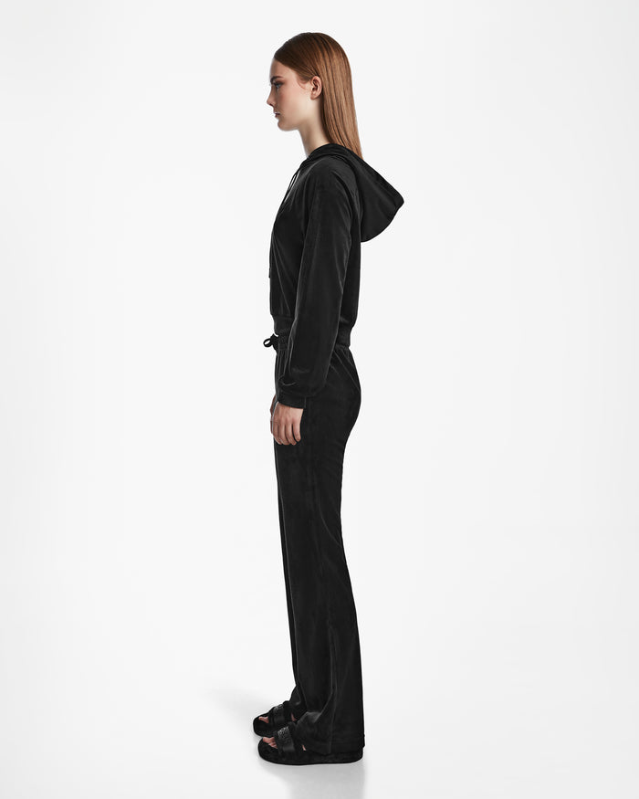 A woman in profile faces left, modeling the Rudsak JODIE - L523501 Black hooded tracksuit with an elastic waistband and straight-leg fit, paired with black sandals, set against a plain white background.