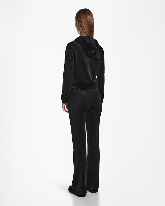 A person with long straight brown hair stands facing away, wearing the Rudsak JODIE - L523501 Black velour tracksuit with hood, long sleeves, and women’s straight-leg pants against a plain white background.