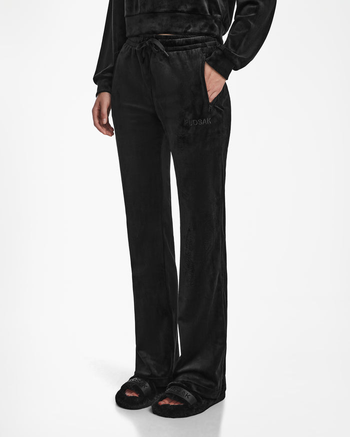 A person wears the Rudsak JODIE - L523501 Black velour lounge pants with an elastic waistband, straight-leg silhouette, matching long-sleeve top, and black fuzzy slide slippers. This cozy set stands out against a white background.