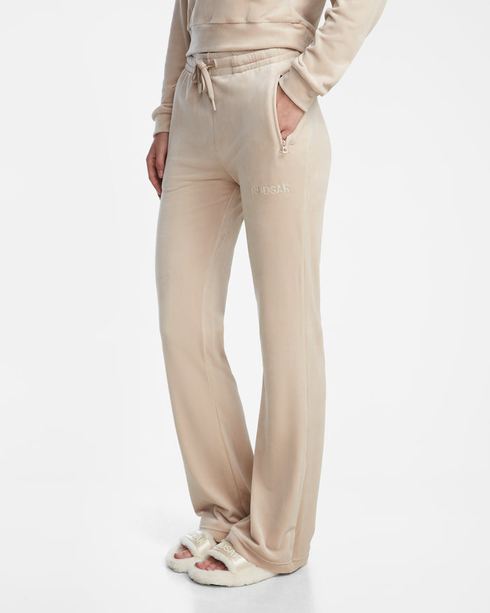 Wearing the Rudsak JODIE - L523501 Cream velour sweatpants and matching hoodie with hands in pockets, paired with fuzzy white slippers, a person stands against a plain white background.
