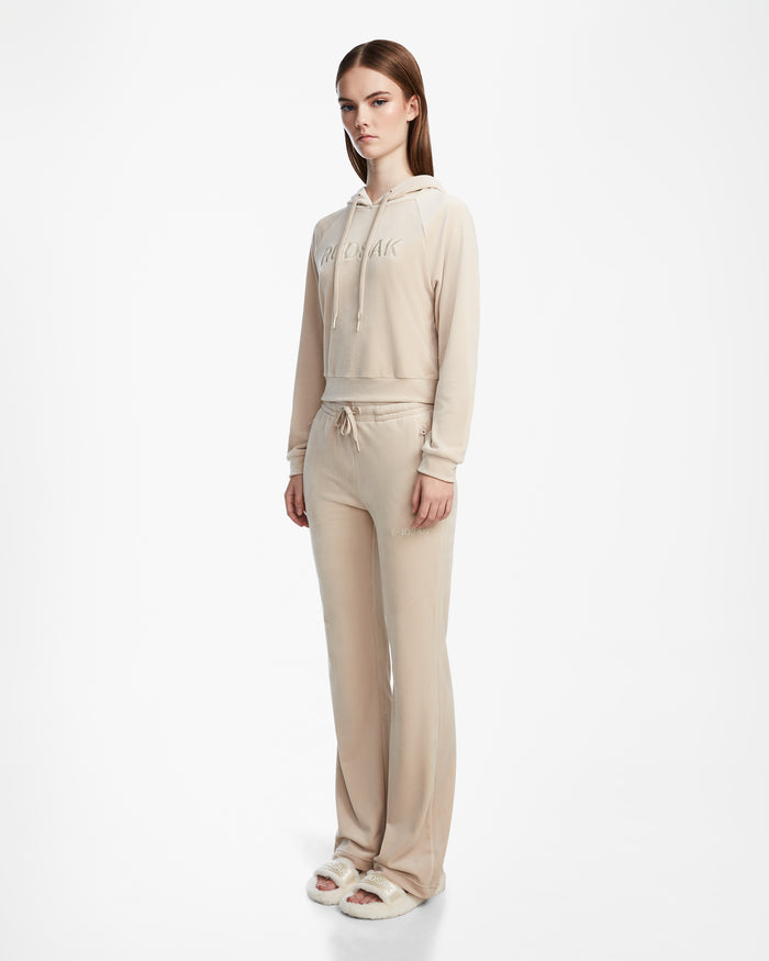 A woman with straight brown hair and a neutral expression stands against a white background wearing the Rudsak JODIE - L523501 Cream velour hoodie.