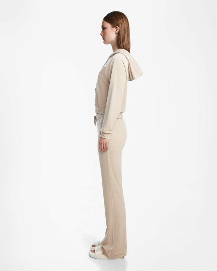 A woman in profile poses against a white backdrop, wearing the Rudsak JODIE - L523501 Cream hooded jacket with straight-leg elastic-waist pants and white sandals. She has straight, long hair and a neutral expression.
