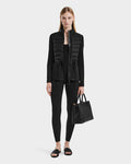 A woman with straight, center-parted blonde hair stands against a plain background wearing the JOSEPHINE - L125011 Black vegan leather quilted jacket by Rudsak, paired with black leggings, slide sandals, and holding a black tote bag.