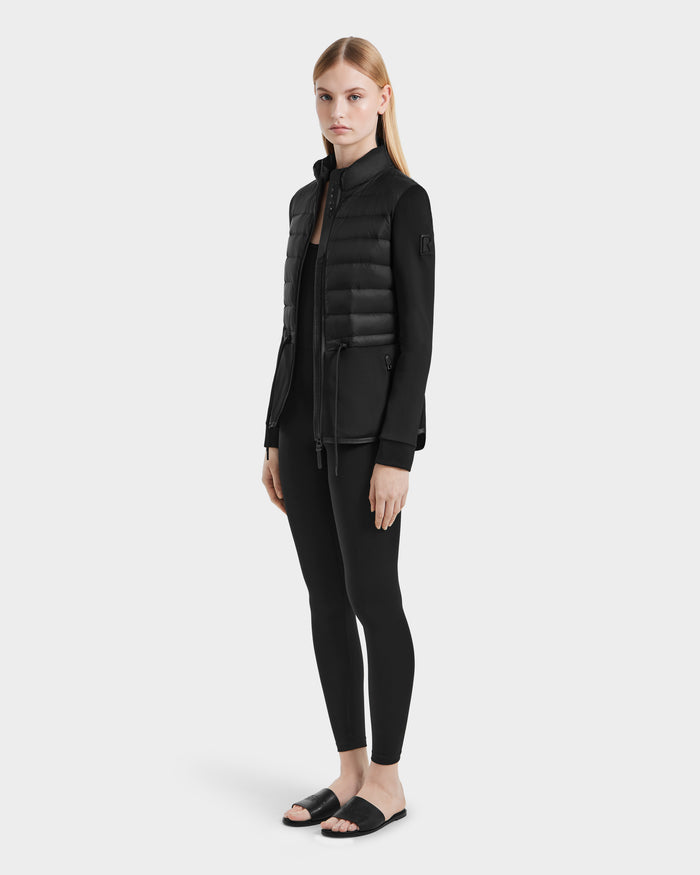 A woman with straight blonde hair and a neutral expression wears the Rudsak JOSEPHINE - L125011 Black vegan leather quilted jacket, black leggings, and slide sandals against a plain background.