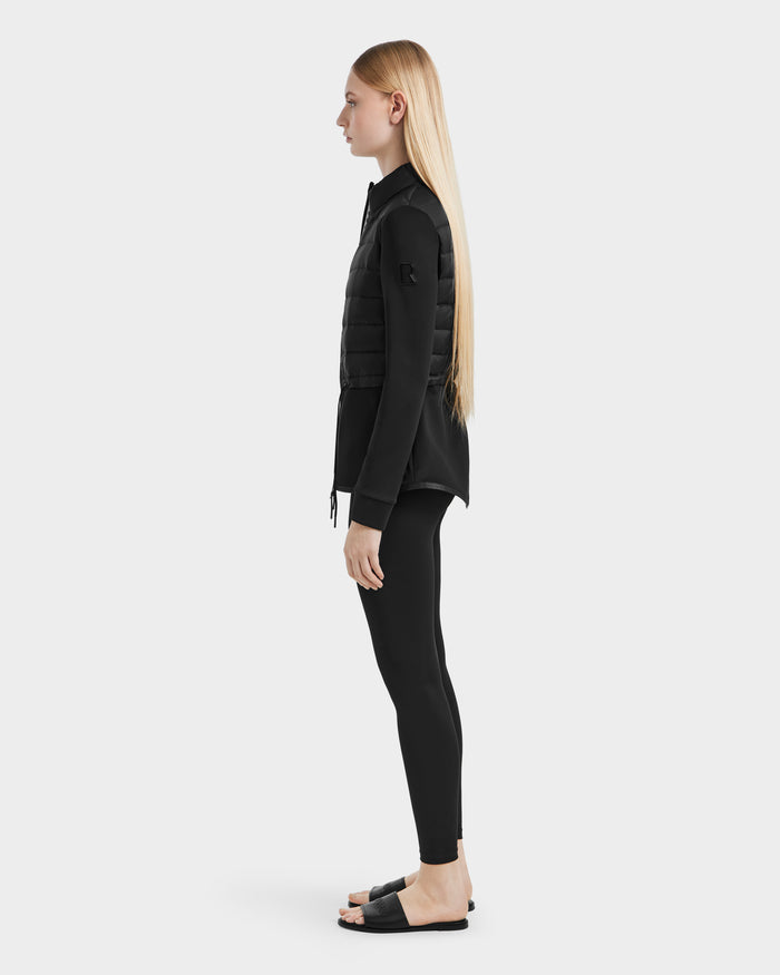 A woman with long blonde hair stands in profile against a plain white background, wearing the JOSEPHINE - L125011 Black hybrid jacket by Rudsak, paired with black leggings and black slide sandals.