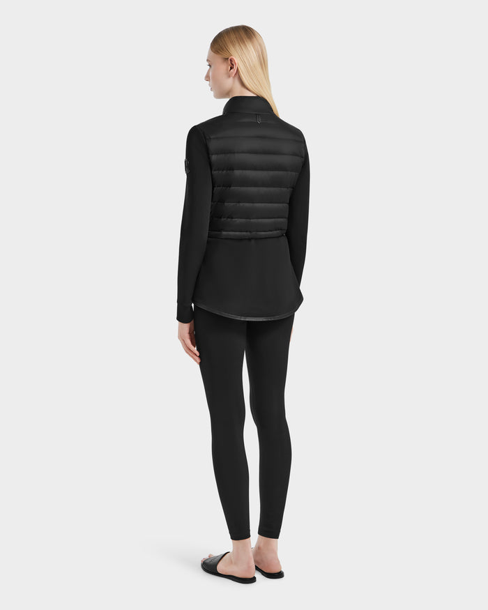 A person with straight blonde hair faces away, wearing the Rudsak JOSEPHINE - L125011 Black recycled nylon puffer vest over a black long-sleeve top, black leggings, and black slide sandals against a plain light background.