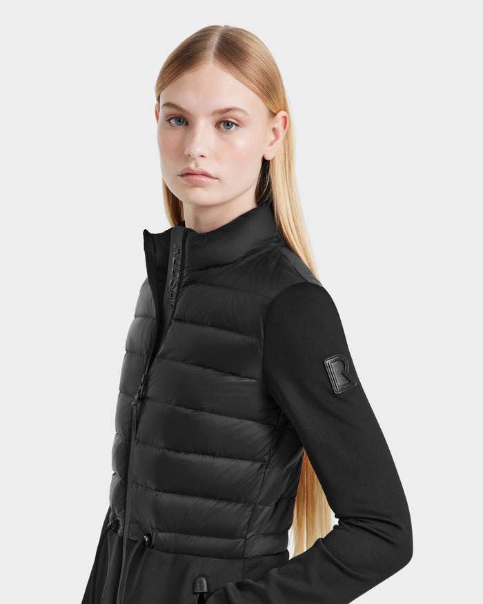 A young woman with straight blonde hair wears the Rudsak JOSEPHINE - L125011 Black quilted hybrid jacket, made from recycled nylon with a zip-up high collar and “R” logo patch on the sleeve, standing against a light gray background.
