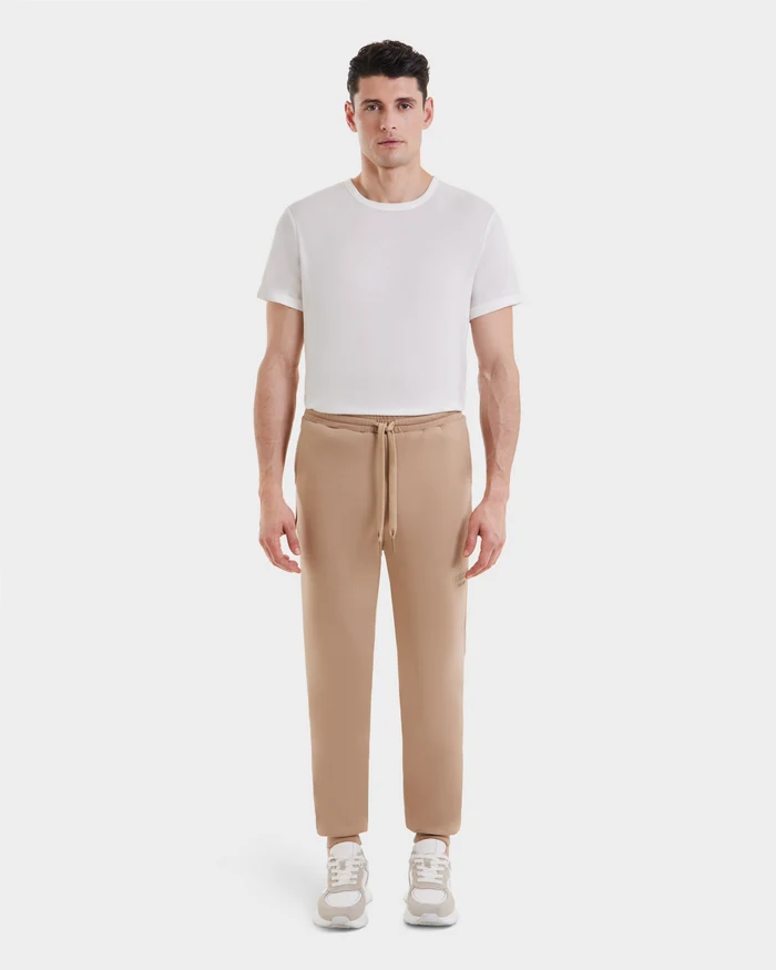 A man stands facing forward against a white background, wearing Rudsak JOSS - 9524009 Almond M premium cotton joggers with a plain white T-shirt and beige sneakers.