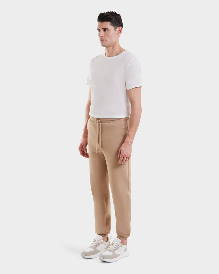 A man stands against a plain white background, wearing the Rudsak JOSS - 9524009 Almond M premium cotton joggers with a drawstring, paired with a white short-sleeve t-shirt and white sneakers.