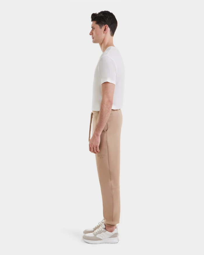 A man stands in profile against a white background, wearing a white t-shirt, Rudsak joggers (JOSS - 9524009 Almond M), and beige-and-white sneakers.
