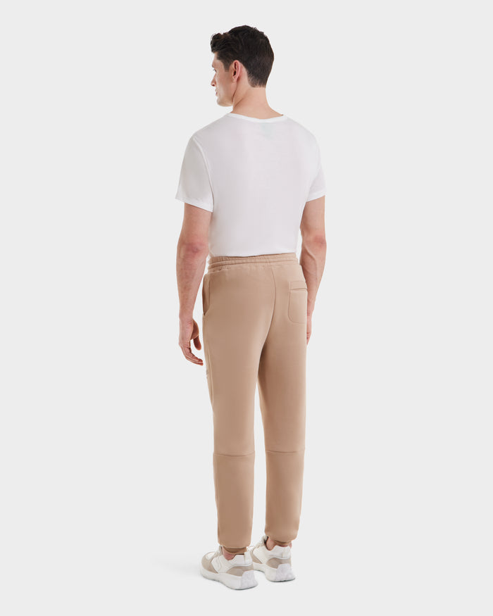 A man stands facing away, wearing Rudsaks JOSS - 9524009 Almond M jogger pants with a white t-shirt and sneakers, set against a plain white background.