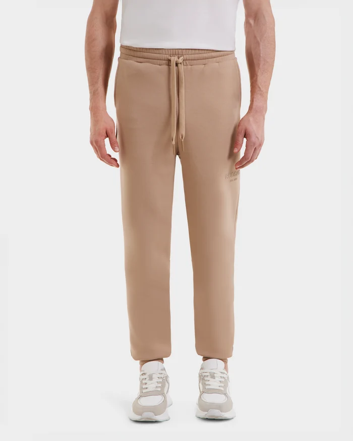 A person wears Rudsaks JOSS - 9524009 Almond M joggers in light beige with a white shirt and sneakers, standing against a plain white background. These premium cotton unisex joggers offer a relaxed fit and drawstring waist for comfort.