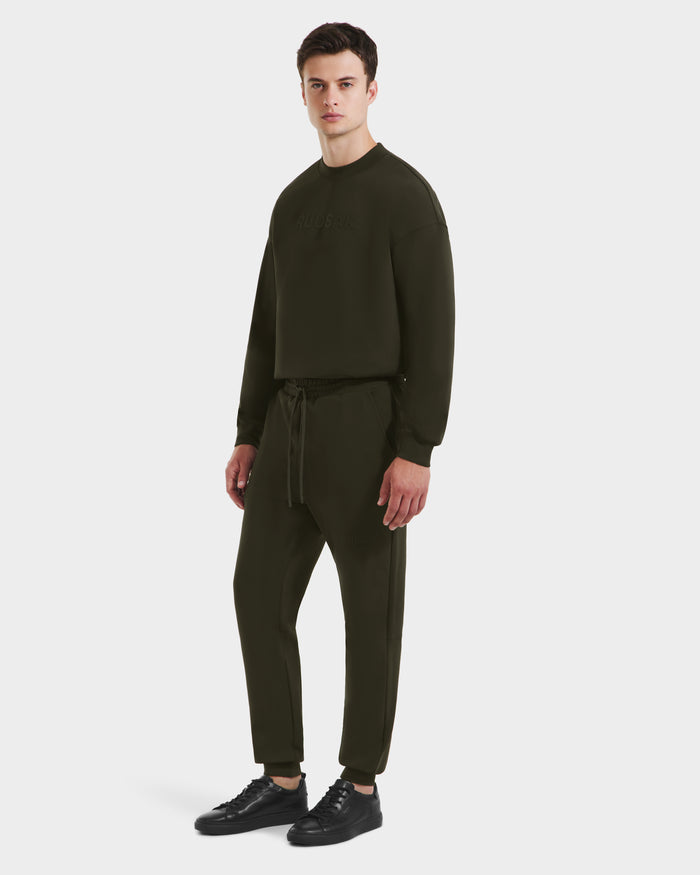 A young man stands against a white background, showcasing the Rudsak JOSS - 9524009 joggers in dark olive (M), paired with a matching sweatshirt and black sneakers, embodying a relaxed modern jogger style.