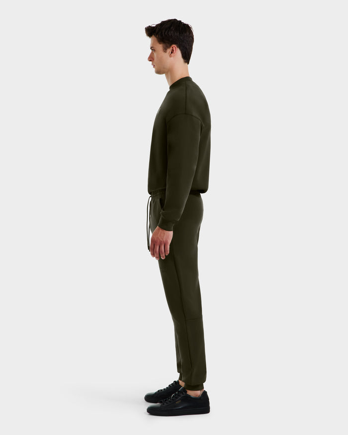A man in profile faces left, wearing the Rudsak JOSS - 9524009 Dark Olive M jogger set with black sneakers, posed against a plain light gray background.