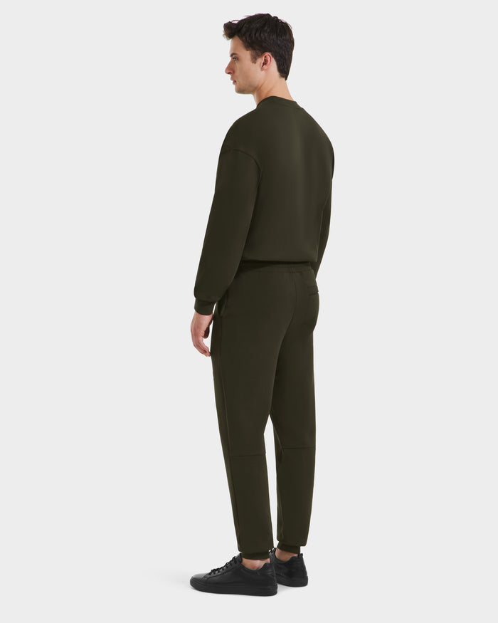 A man faces slightly away, wearing the Rudsak JOSS - 9524009 jogger sweatshirt and sweatpants set in Dark Olive (size M) with black sneakers, against a plain white background.