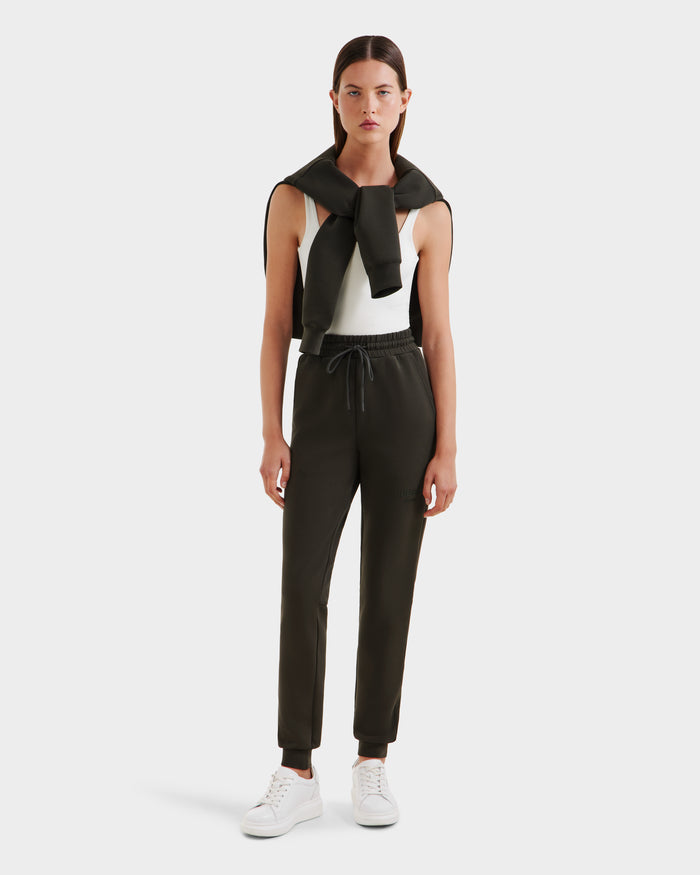 A woman stands facing forward in Rudsaks JOSS - 9524009 Dark Olive W joggers, white sleeveless top, white sneakers, and a black sweater draped over her shoulders, set against a plain white background.