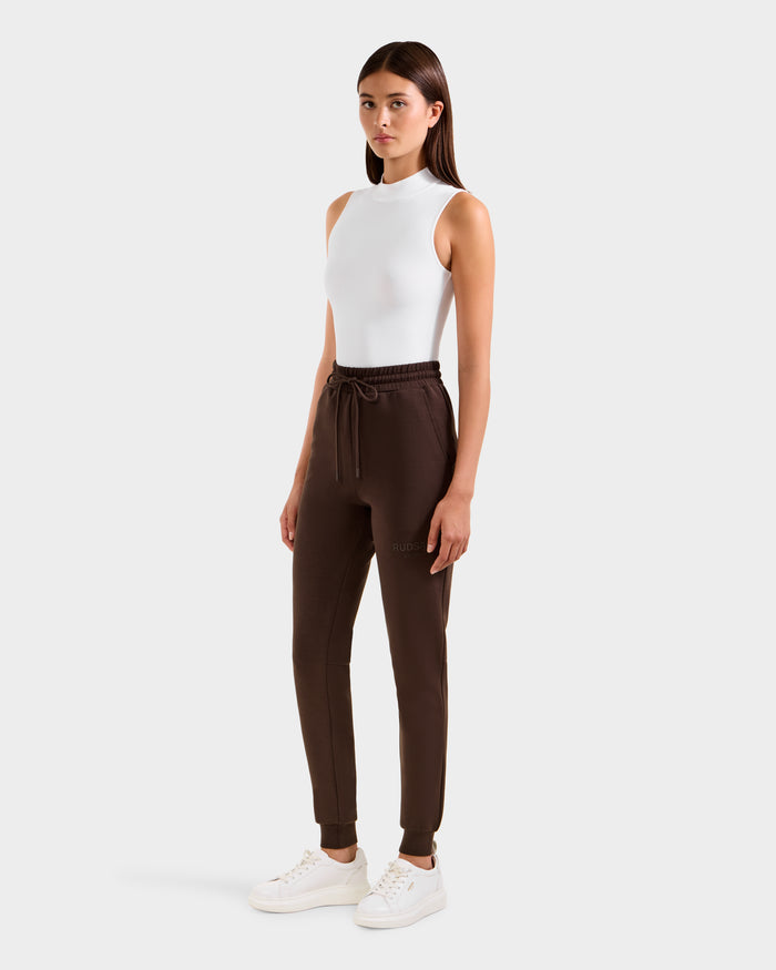 A woman stands facing forward wearing Rudsak's JOSS - 9524009 Espresso W modern jogger dark brown drawstring pants, styled with a sleeveless white top and white sneakers against a plain light background.