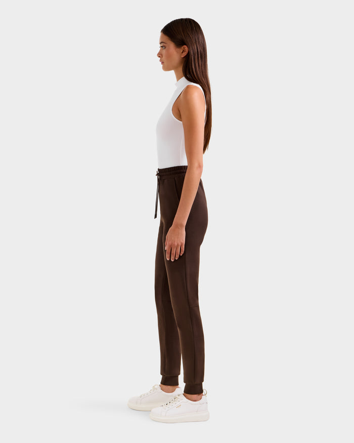 A woman stands in profile against a plain background, wearing Rudsak's JOSS - 9524009 Espresso W premium cotton jogger pants in brown, paired with a white sleeveless top and white sneakers. Her long hair is straight and she looks ahead, arms relaxed.