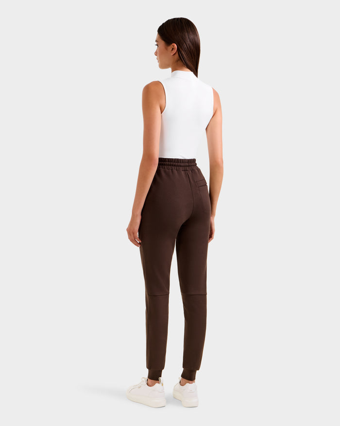 A woman stands facing away, wearing Rudsak's JOSS - 9524009 Espresso W premium cotton jogger pants in brown, paired with a sleeveless white top and white sneakers against a plain light gray background.
