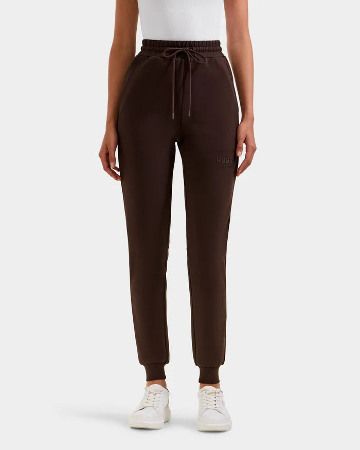 Wearing Rudsak’s JOSS - 9524009 Espresso W joggers, known for their unisex fit, drawstring waist, and cuffed ankles, paired with a white sleeveless top and sneakers, a person stands against a plain light background.