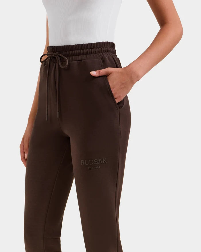 A person wears Rudsak’s JOSS - 9524009 Espresso W unisex joggers in dark brown premium cotton with an elastic waistband, drawstring, pockets, and “RUDSAK EST. 1994” embroidered on the upper thigh.