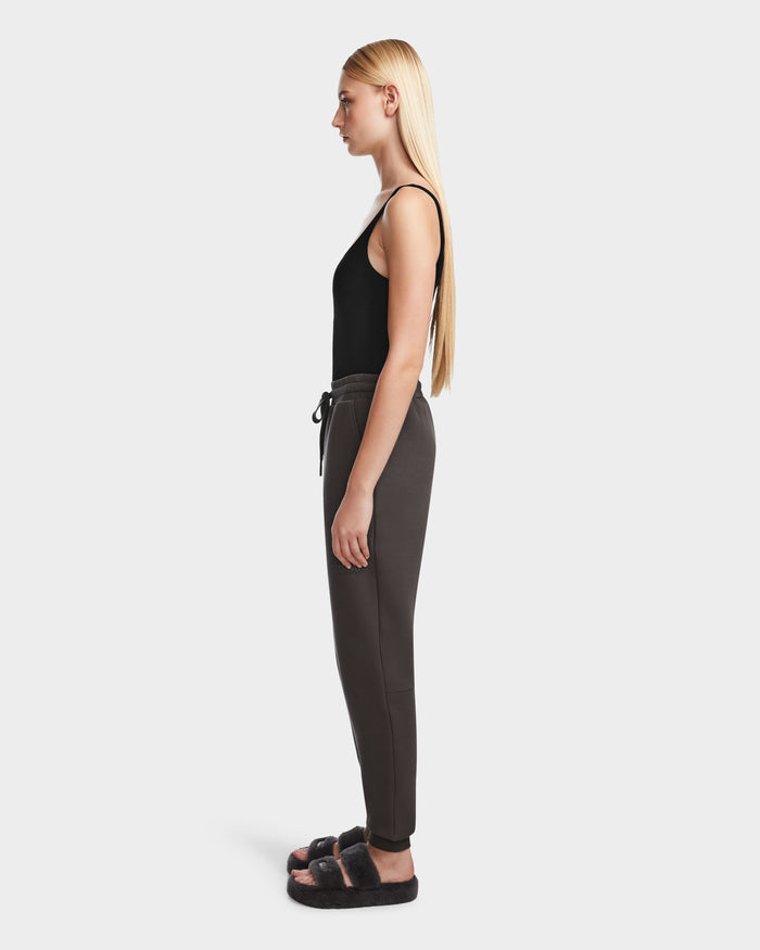 A woman with long blonde hair stands in profile against a white background, wearing a black sleeveless top, Rudsak JOSS - 9524009 Grey W premium cotton joggers with a drawstring, and black fuzzy slide sandals.