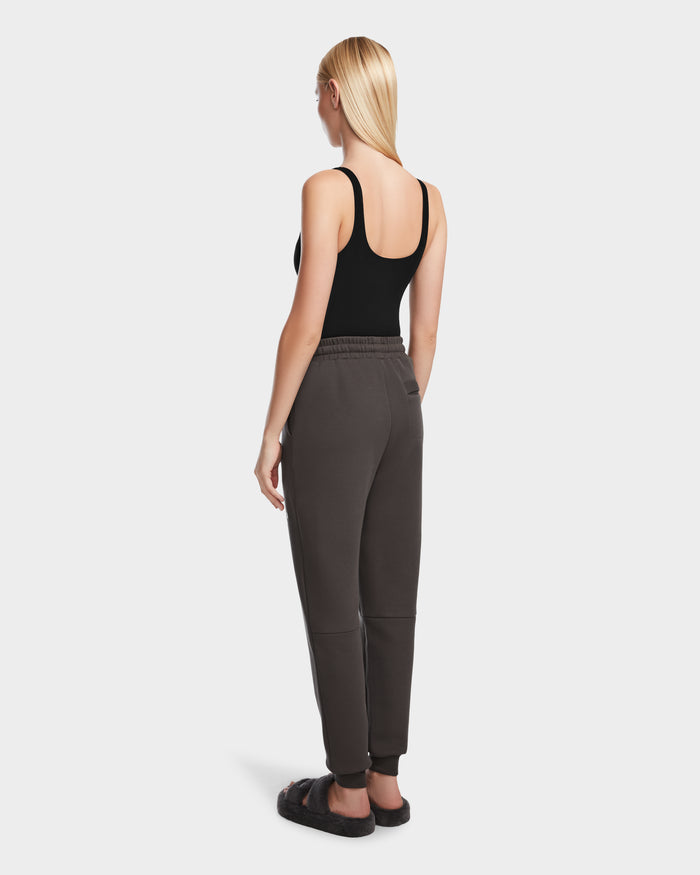 A woman with straight blonde hair, wearing a black tank top and Rudsak JOSS - 9524009 Grey W premium cotton joggers in dark gray, stands facing away from the camera on a plain white background.