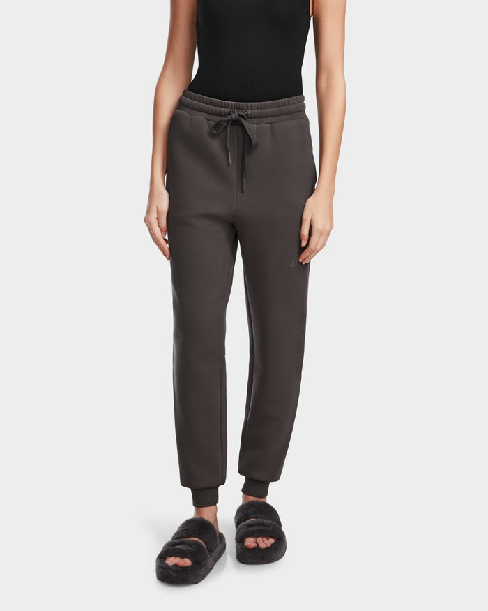A woman wears the Rudsak JOSS - 9524009 Grey W joggers with a drawstring, styled with a black sleeveless top and fuzzy slippers. She stands facing forward, arms relaxed at her sides against a plain background.