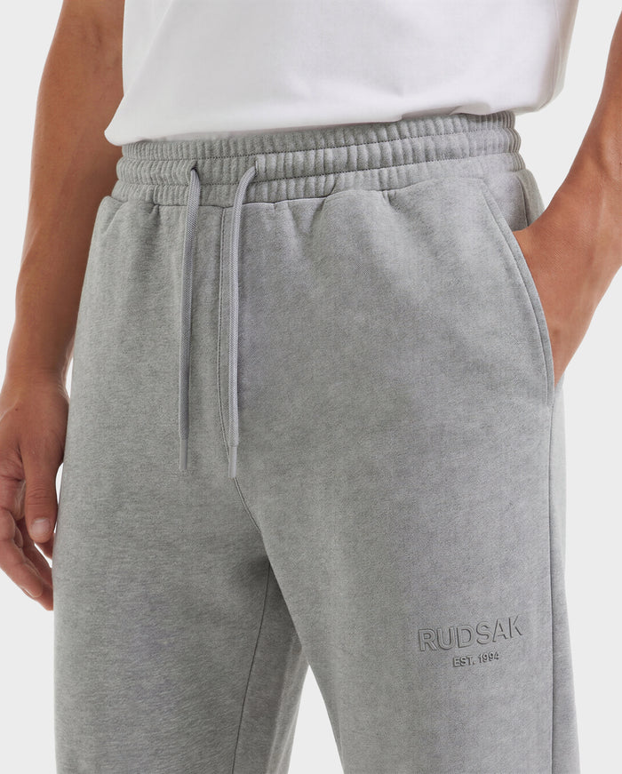 A person wears Rudsak JOSS - 9524009 joggers in heather grey (size M) with an elastic waistband, drawstrings, and "RUDSAK EST. 1994" on the left thigh, paired with a white shirt and their left hand in the pocket.