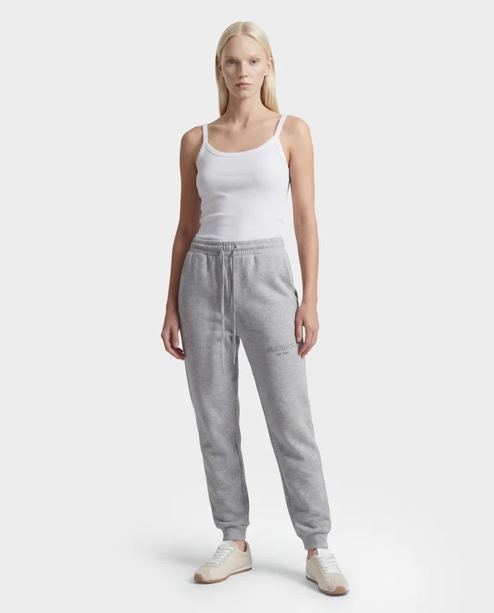 A woman stands facing forward against a plain white background, wearing the Rudsak JOSS - 9524009 Heather Grey W joggers, a white tank top, and white sneakers.