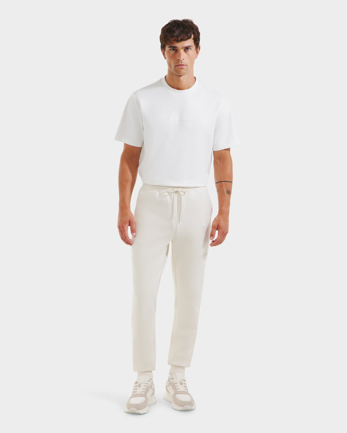 A man faces forward against a plain light background, wearing the Rudsak JOSS - 9524009 Vanilla M premium cotton joggers in cream, a white short-sleeve t-shirt, and beige-white sneakers.