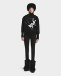 A woman faces forward in the Rudsak JOYCE - 8525647 Black sweater, featuring a chic white abstract skiing graphic. She pairs it with black leggings, a furry hat, and matching boots against a plain white background.