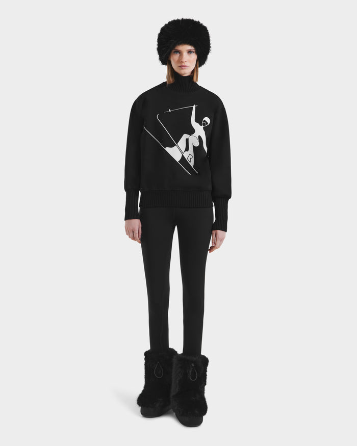 A woman faces forward in the Rudsak JOYCE - 8525647 Black sweater, featuring a chic white abstract skiing graphic. She pairs it with black leggings, a furry hat, and matching boots against a plain white background.