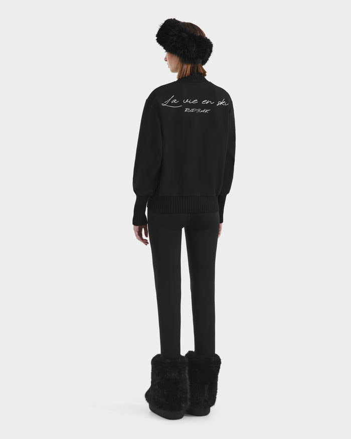 A person stands facing away, wearing the Rudsak JOYCE - 8525647 Black après-ski sweater with fur boots and a fur headband. The back of the sweater features white script. The background is plain white.