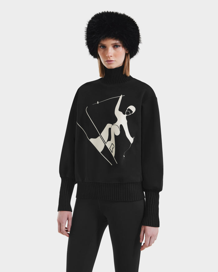 A woman models the Rudsak JOYCE - 8525647 Black turtleneck, featuring an abstract white skiing figure, with black pants and a fur hat against a plain white backdrop.