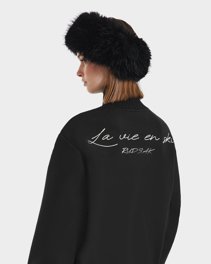 A woman is shown from the back in side profile, wearing the Rudsak JOYCE - 8525647 Black furry headband and a womens ski sweater with “La vie en ski” and “BUNSHAK” text against a plain light background.