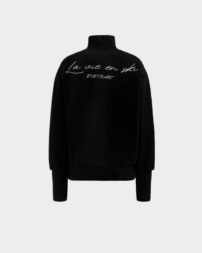 The Rudsak JOYCE - 8525647 Black women’s ski sweater features ribbed cuffs and hem, plus white cursive "La vie en ski" and "NDSAK" on the back—perfect for après-ski style against a light gray background.