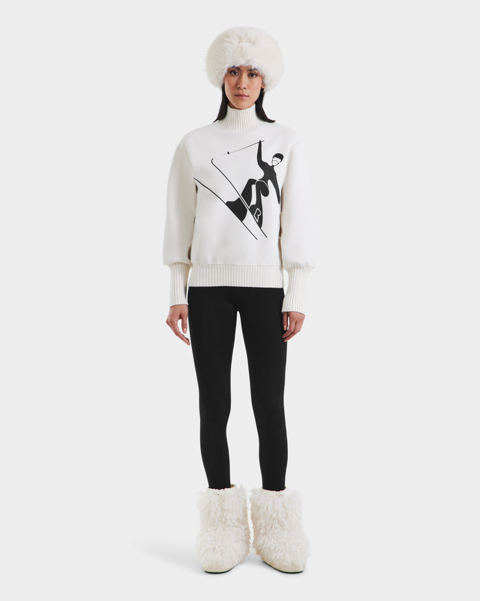 A person faces forward against a plain background, dressed in the Rudsak JOYCE - 8525647 Vanilla vintage ski sweater with a black skiing graphic, paired with black leggings, fluffy white boots, and a furry white hat.