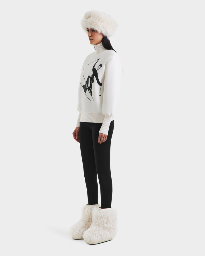 A woman stands sideways wearing the Rudsak JOYCE - 8525647 Vanilla women’s ski sweater, featuring a black abstract design, paired with black leggings, a white furry hat, and large fluffy boots against a plain background.