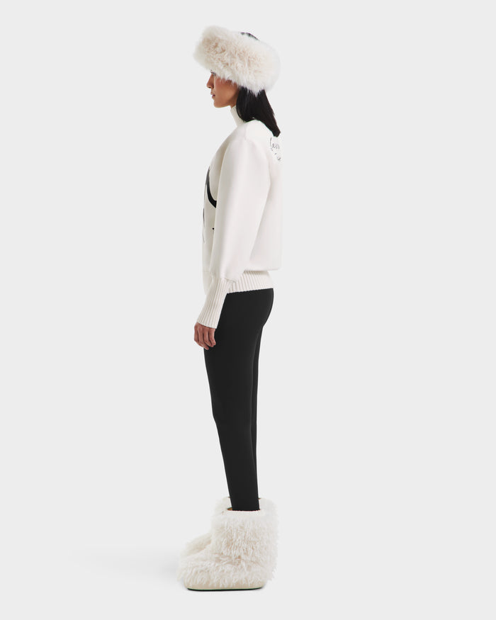 A woman in profile wears the Rudsak JOYCE - 8525647 Vanilla white fur hat, après-ski sweater, white jacket, black leggings, and large white fur boots against a plain white background.