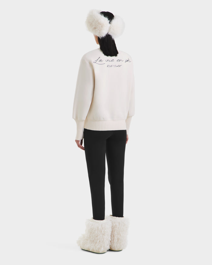 A person faces away, wearing the Rudsak JOYCE - 8525647 Vanilla retro ski sweater with black pants, white earmuffs, and matching fluffy boots, all set against a plain white background.