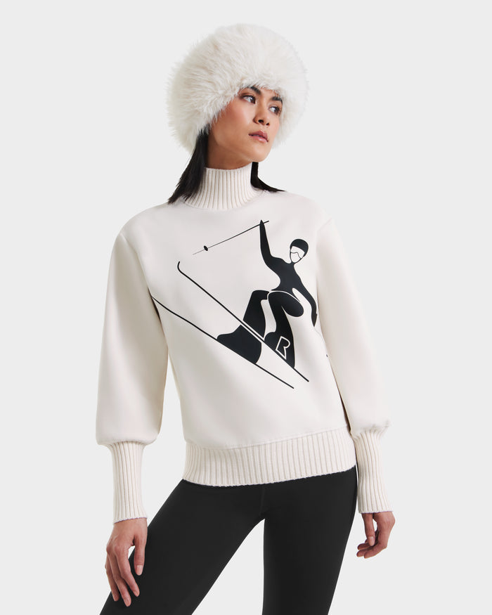 A woman models the Rudsak JOYCE - 8525647 Vanilla sweater featuring a black skier graphic, styled with a white fur hat and black pants against a plain white background.