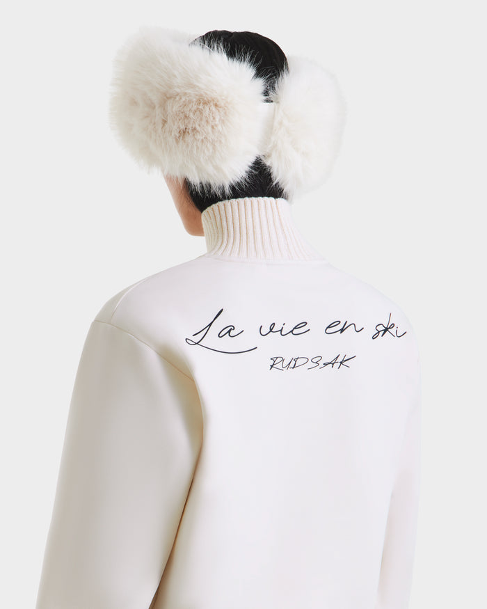 A person with short dark hair wears large white fur earmuffs and the Rudsak JOYCE - 8525647 Vanilla retro ski sweater jacket with “La vie en ski” in black cursive on the back, against a plain white background.