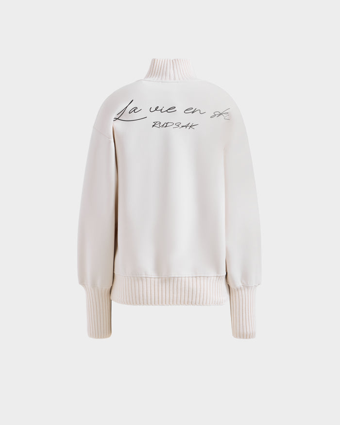 The JOYCE - 8525647 Vanilla by Rudsak is a cream women’s long-sleeve ski sweater with ribbed cuffs and hem, featuring "La vie en rose" and "BADSAK" embroidered in black script on the back.