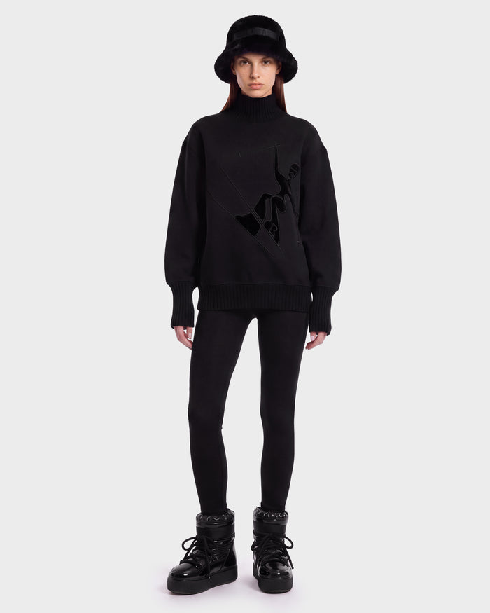 A woman wearing the Rudsak JOYCE E - 8526538 Black bucket hat stands against a plain white background, styled with an oversized black turtleneck sweater featuring a graphic design, black leggings, and black winter boots.