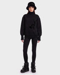A woman wearing the Rudsak JOYCE E - 8526538 Black bucket hat stands against a plain white background, styled with an oversized black turtleneck sweater featuring a graphic design, black leggings, and black winter boots.
