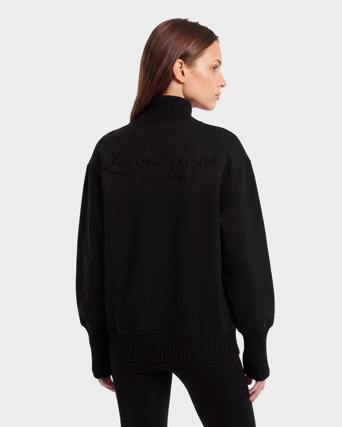A woman with straight brown hair wears the Rudsak JOYCE E - 8526538 Black turtleneck sweater, featuring embossed text on the back, standing against a plain light gray background.
